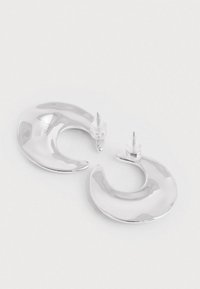 O pereche de cercei hoop în formă de semilună, argintii și strălucitori, cu suporturi transparente, așezați pe o suprafață albă simplă.