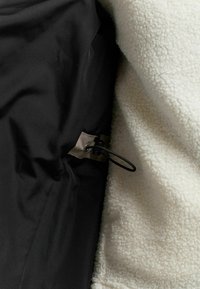 Musta ulkokerros, jossa on sileä pinta ja säädettävä naru, sekä kermaisen värinen, pörröinen fleecevuori, jossa on pehmeä ja teksturoitu pinta.