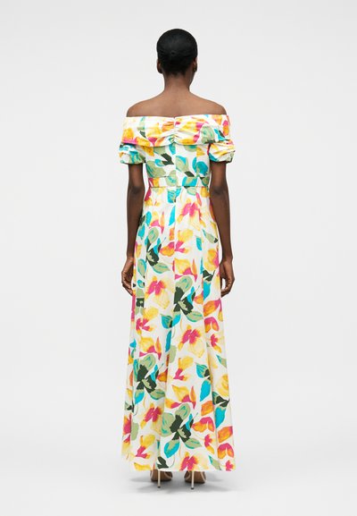 Sieviete redzama no aizmugures, valkājot baltu, līdz zemei krītošu kleitu bez plecu daļas, ar krāsainu ziedu rakstu un īsām pūkainām piedurknēm.