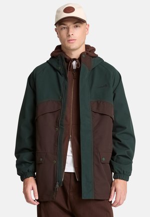 Young man wearing a green and brown hooded jacket over a brown zip-up hoodie, a white cap with a brown patch, and brown pants.