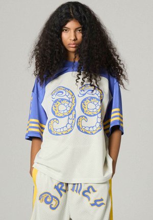 Młoda osoba z długimi kręconymi włosami, ubrana w luźne białe i niebieskie ubranie sportowe z żółto-niebieskimi wzorami graficznymi oraz numerem 99.