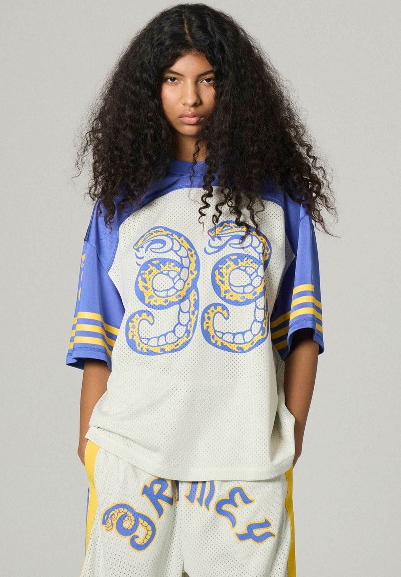 Persona joven con cabello largo y rizado que lleva ropa deportiva suelta blanca y azul con diseños gráficos en amarillo y azul y el número 99.