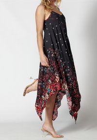 Femme portant une robe noire à ourlet asymétrique fleuri et des sandales à talons transparents, debout avec un pied levé derrière elle.