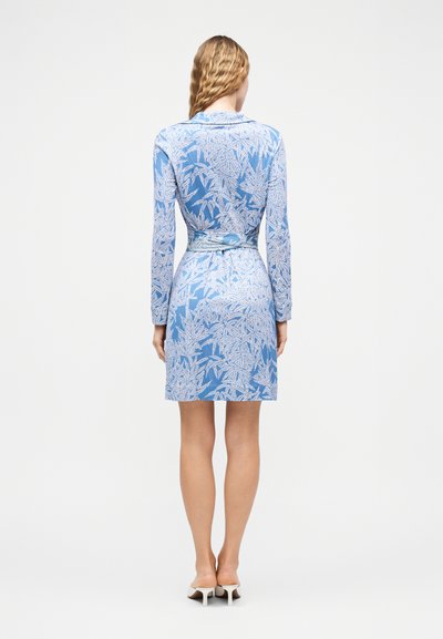 Γυναίκα με κυματιστά μαλλιά φοράει ένα ανοιχτό μπλε μίνι φόρεμα με λευκό φύλλωμα, μακριά μανίκια και λευκά mule με τακούνι, στέκεται γυρισμένη προς τα πίσω.