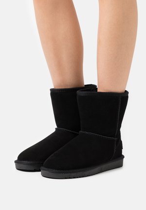 Piernas vestidas con botas de ante negras hasta la mitad de la pantorrilla, con suelas de goma planas, costuras visibles y punteras redondeadas, frente a un fondo blanco liso.