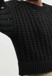 Nahaufnahme einer Person, die einen schwarzen, gestrickten Pullover mit senkrechten, offenen Webmustern sowie einem gerippten Saum und Halsausschnitt trägt.