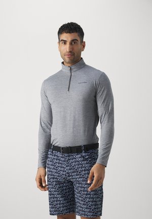 Grå langermet zip-up skjorte med høy krage, med en liten svart logo på brystet, kombinert med marineblå shorts med hvit tekst.