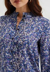 Chemise à motifs floraux avec un motif violet et noir, dotée d'un col froncé et de boutons noirs à l'avant. Texture du tissu douce.