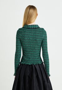 Groen geruite, nauwsluitende top met lange mouwen, textuurstof; gerimpelde zoom, contrasterende kraag. Draagt met een zwarte, volumineuze rok.