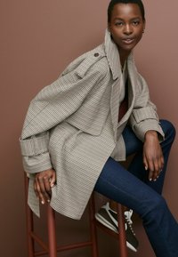 Ruuduline trench coat beeži ja musta värviga, suure kaelusega ja nööbiga detailidega. Koos tumedate teksade ja hallide sneakeritega. Lihtne taust.