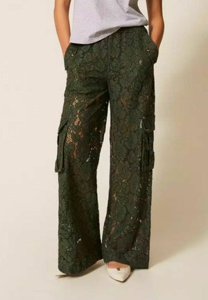 Pantaloni cargo in pizzo verde scuro a gamba larga, indossati con scarpe bianche appuntite e top grigio chiaro, mani nelle tasche.