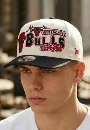 Junger Mann, der eine weiße und schwarze Chicago Bulls-Kappe mit den Aufschriften "1966" und Bull-Logos trägt und leicht nach unten blickt, im Freien.