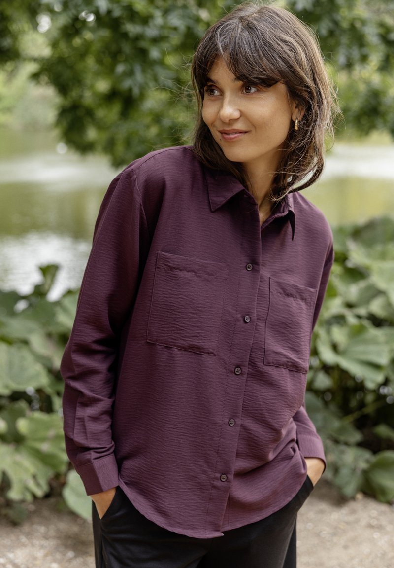 Camicia button-up viola realizzata in tessuto testurizzato, con colletto e due tasche sul petto. Indossata su pantaloni neri in un contesto naturale.