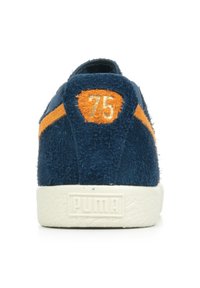 Baskets en suede bleu marine avec une rayure orange et un emblème « 75 » sur le talon. Semelle en caoutchouc texturé blanc avec la marque PUMA.