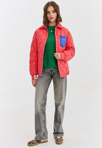 Žena v korálové prošívané bundě s modrým kapesníkem přes zelený svetr, šedé džíny a boty s leopardím vzorem stojí na bílém pozadí.