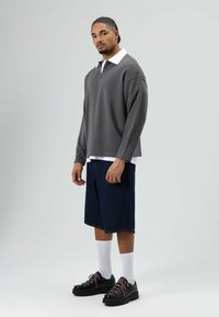 Grijze sweater met ritssluiting aan de voorkant en een witte kraag, gecombineerd met donkere spijkershorts. Zwarte schoenen met roze veters en witte sokken maken de outfit compleet.