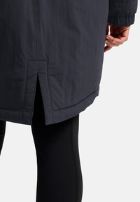 Waterdichte, donkermarine jas met een rechte zoom en zijsplit, met een gladde textuur en elastische manchetten. Draagt over zwarte leggings.