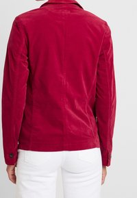 Henkilö, joka on pukeutunut istuvaan pitkähihaiseen punaiseen takkiin, jossa on napitettu hihansuu, ja valkoisiin housuihin. Henkilö on kuvattu selkäpuolelta yksinkertaista taustaa vasten.