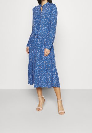 Vestido azul de manga larga con un ligero cuello, patrón floral en tonos más claros, cintura fruncida y falda fluida. Combinado con tacones color beige pálido.