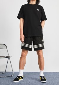 Černé bavlněné tričko s malým bílým logem, spárované s černými šortkami s listovým vzorem a bílými pruhovanými akcenty. Černé a žluté tenisky.