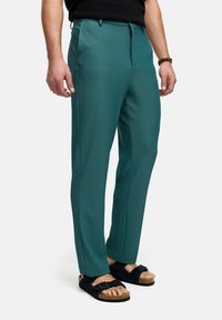 Teal plisserede bukser med en skræddersyet pasform, der har lynlås lukning og bæltestropper, parret med sorte sandaler på bare fødder.