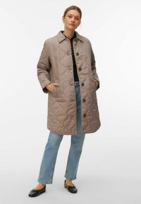 Naine seisab käed taskutes, seljas põlveni ulatuv tuhmunud beež ruuduline mantel, helesinised teksad, must top ja mustad baleriinikingad.