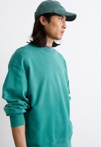 Tyrkis sweatshirt med rund halsudskæring og ribbede manchetter, lavet af blødt stof. Parret med en matchende tyrkis kordfløjlskasket.