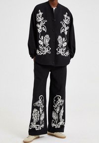 Chaqueta negra con bordados florales blancos y botones, combinada con pantalones negros a juego que presentan patrones detallados similares. Tejido texturizado.