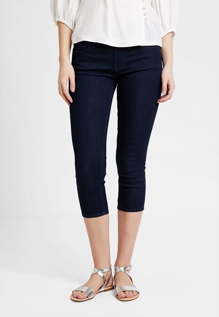 Mujer con pantalones pitillo cortos de color azul oscuro, blusa de textura blanca y sandalias planas plateadas con tiras en el tobillo.