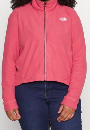 Kvinde iført en knaldrød North Face fleecejakke med lynlås, kombineret med mørkeblå jeans, mod en ensfarvet baggrund.