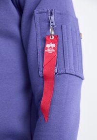 Felpa viola con zip e un'etichetta di stoffa con un nastro rosso, che presenta il logo "Alpha Industries". Morbido al tatto e con tasche.