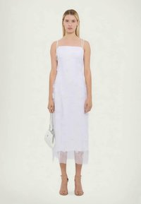 Λευκό αμάνικο φόρεμα με λεπτομέρειες από δαντέλα, ίσια σιλουέτα και ημιδιαφανή κάτω κόψιμο. Συνδυάζεται με μπεζ σανδάλια με λουριά και μια μικρή τσάντα χεριού.