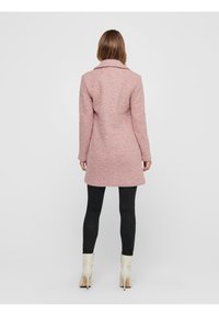 Figurerad rosa kappa i texturerad tyg, med snibbiga kragar och långa ärmar, parat med svarta smala byxor och krämfärgade ankelskor.