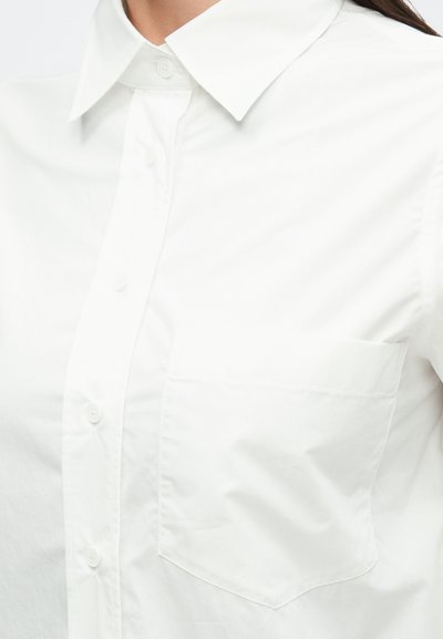 Biela košeľa na gombíky s ostrým golierom, jedným náprsným vreckom a hladkou bavlnenou textúrou. Čistý dizajn, bez vzorov.