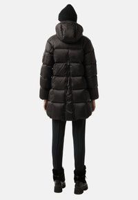 Přefukovací bunda v tmavě černém materiálu, se vzorem jemné quiltování, kapucí a logem na zadní straně. Nosit s přiléhavými černými kalhotami a kotníčkovými botami.