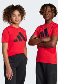 Červené tričká s krátkym rukávom a veľk�ým čiernym logom Adidas. Čierne tepláky s vreckami a decentným brandingom Adidas. Dvaja modeli, stojaci vedľa seba.