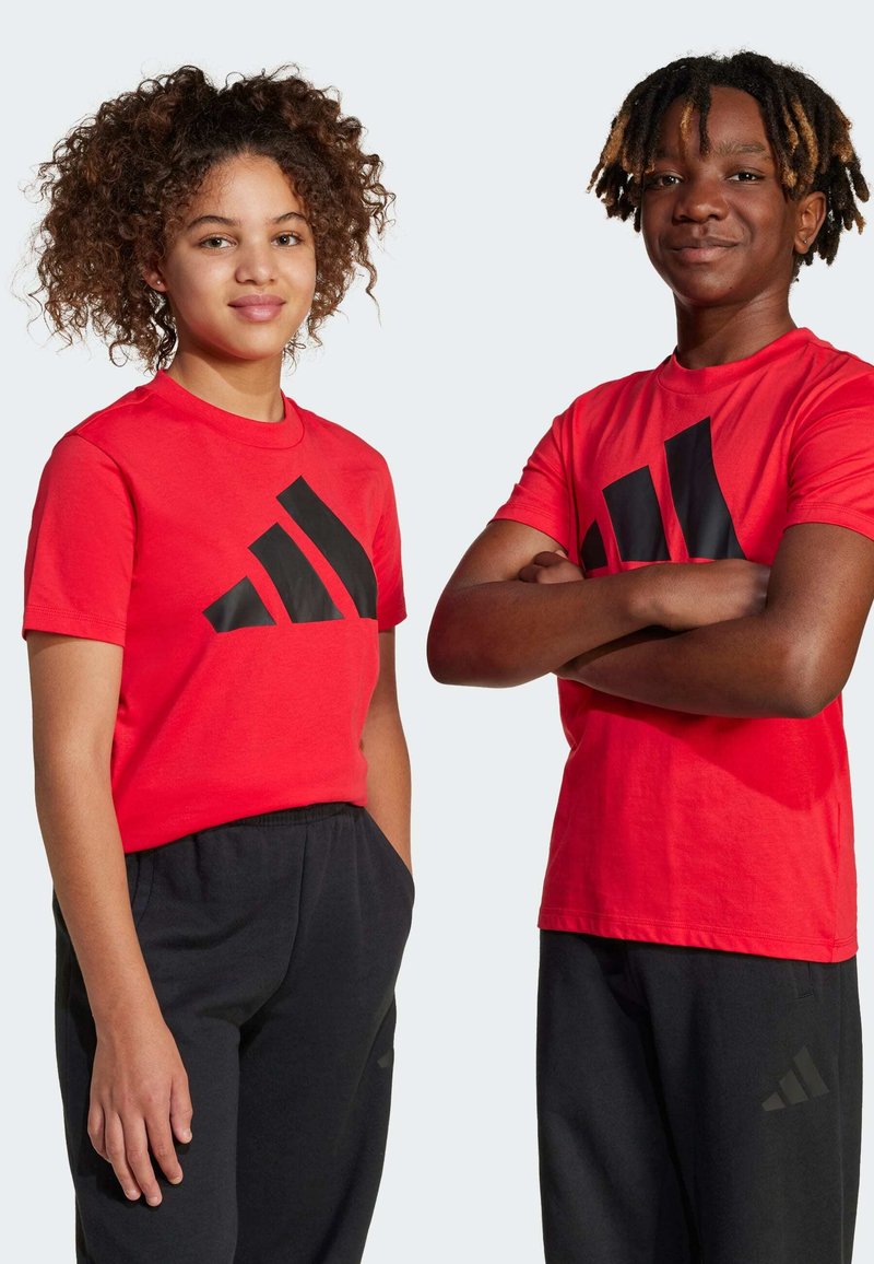 Červené tričká s krátkym rukávom a veľkým čiernym logom Adidas. Čierne tepláky s vreckami a decentným brandingom Adidas. Dvaja modeli, stojaci vedľa seba.