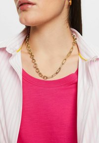 Goldene Kettenhalskette aus ineinandergreifenden Gliedern, getragen über einem pinken Oberteil, kombiniert mit einem weiß-rosa gestreiften Hemd. Glatte, glänzende Oberfläche.