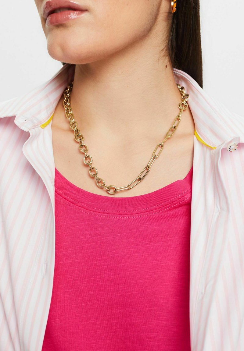 Goldene Kettenhalskette aus ineinandergreifenden Gliedern, getragen über einem pinken Oberteil, kombiniert mit einem weiß-rosa gestreiften Hemd. Glatte, glänzende Oberfläche.