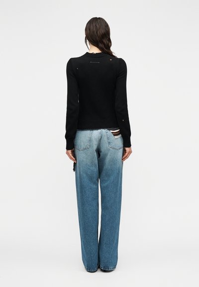 Γυναίκα με μακριά καστανά μαλλιά, που φοράει μαύρο πουλόβερ με φθορές και άνετο ανοιχτό μπλε τζιν, στέκεται μπροστά σε ένα απλό ανοιχτό φόντο.