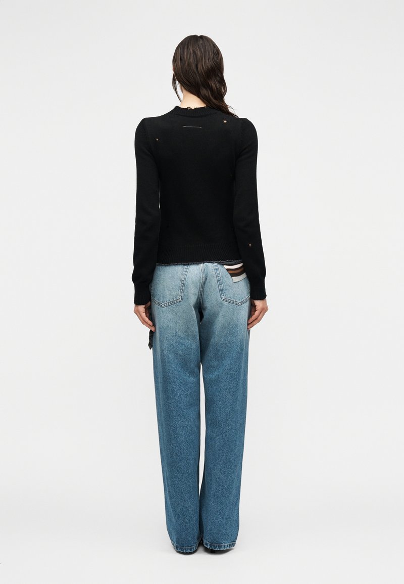 Femme aux longs cheveux bruns portant un pull noir vieilli et un jean large bleu clair, debout devant un fond clair uni.