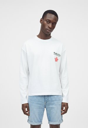 Homme portant une chemise blanche à manches longues avec une étoile rouge et un texte sur la poitrine, associée à un short en denim bleu clair retroussé.