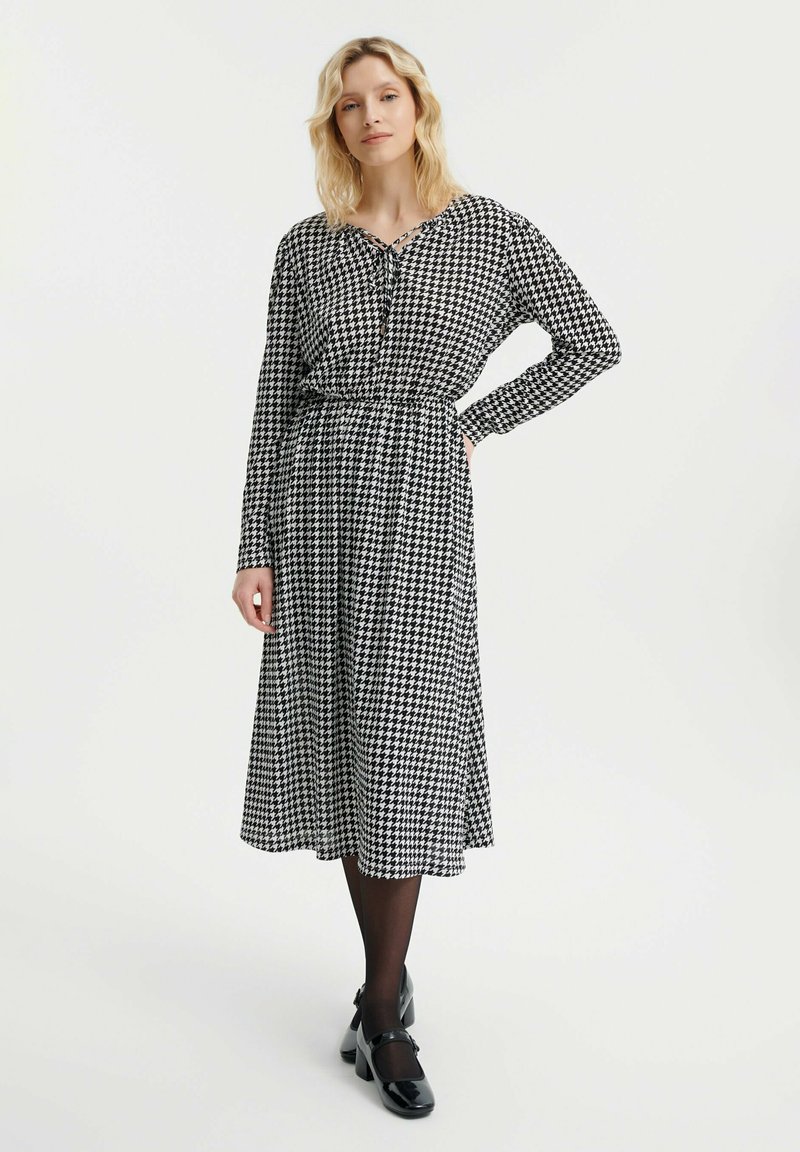Vestido de patrón de pied de poule en blanco y negro con mangas largas, escote atado y silueta en A, combinado con zapatos Mary Jane negros.