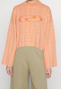 Skrócony, oversize sweter z teksturowej pomarańczowej tkaniny z graficznym wzorem w wir. Posiada wysoki dekolt i szerokie rękawy. Połączony z beżowymi spodniami.