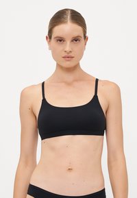 Woman with slicked-back hair wearing a black sports bra and black underwear, facing forward against a plain white background.