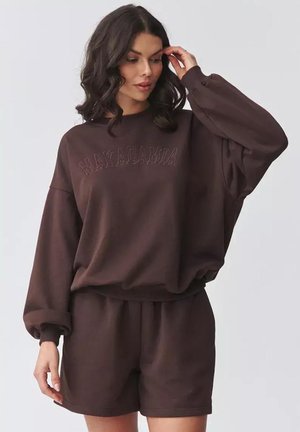 Kobieta z czarnymi, falującymi włosami, ubrana w luźną ciemnobrązową bluzę z napisem "Makadamia" oraz dopasowane szorty, na jednolitym tle.