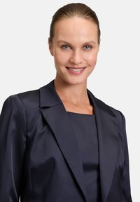 Granatowa marynarka o strukturalnym kroju, satynowej teksturze, klapach w kształcie litery "notch", oraz dopasowanej sylwetce, zestawiona z pasującą ciemną bluzką.