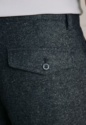 Close-up van donkergrijze textuurstof broek met een klepzak met knopen achterop, gedragen met een wit topje.
