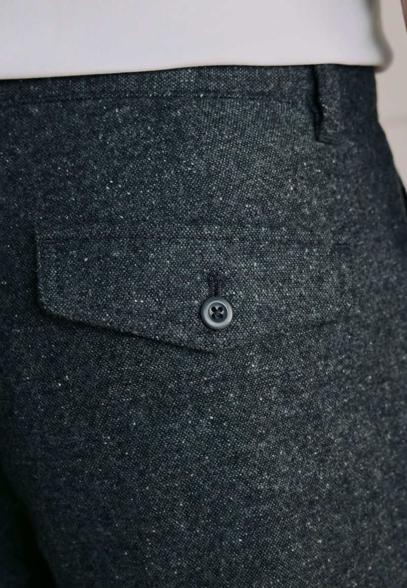 Gros plan d’un pantalon en tissu texturé gris foncé avec une poche à rabat boutonnée à l’arrière, porté avec un haut blanc.