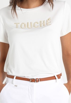 Vrouw draagt een wit T-shirt met "TOUCHÉ" geschreven in parel-besette letters, ingestopt in een witte broek met een bruine riem.
